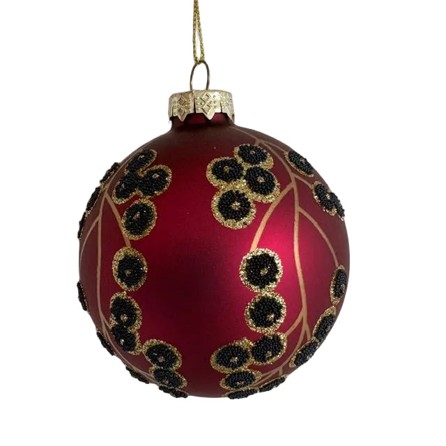 A red glass Christmas ornament with gold glittery branches and black beaded berries, hanging against a plain white wall.