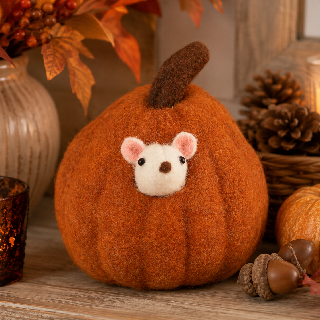 A felted orange pumpkin decoration with a cute white mouse face peeking out, styled on a wooden surface with autumn props including pinecones, acorns, a small pumpkin, and a candle.