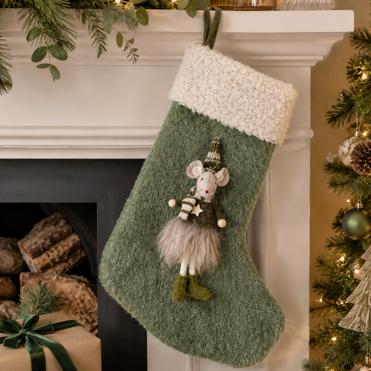 A green textured Christmas stocking with a fluffy white cuff hangs on a white fireplace mantel decorated with pine branches, lights, and wrapped gifts. The stocking features a small mouse figure hanging from the center.